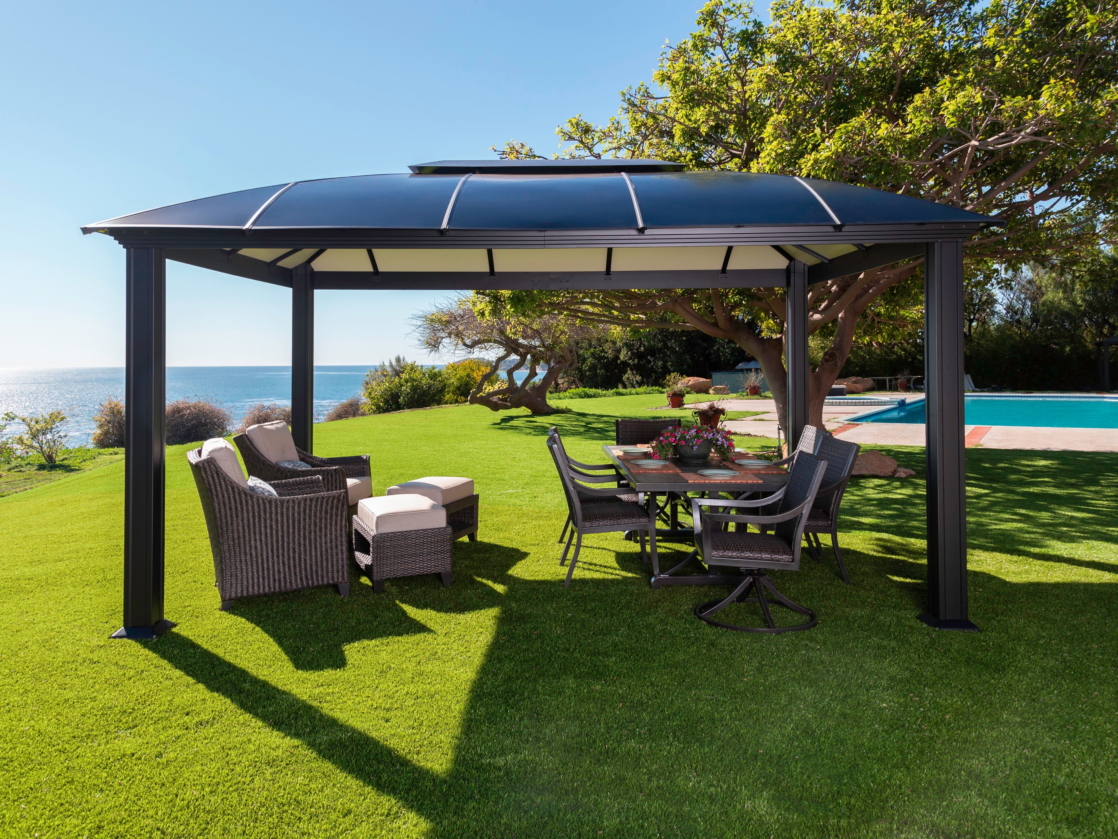 Cambridge 12' x 16' Hard Top Gazebo on grass, in front of pool with outdoor furniture underneath.